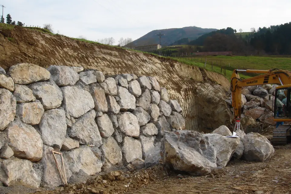 Boulder wall going in at the foot of a cut hillside, with an excavator working nearby