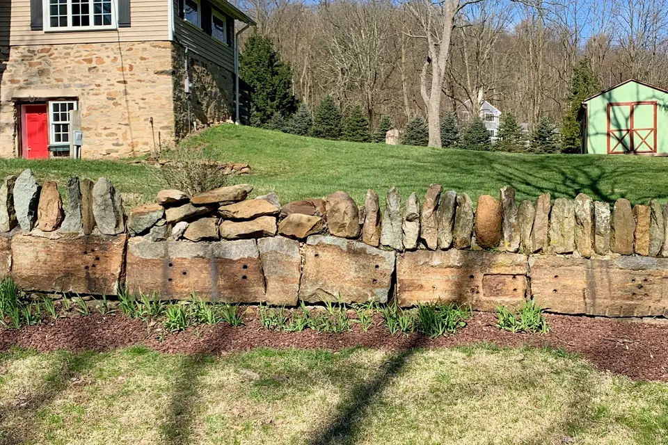 A stacked stone retaining wall edging a sloped lawn and planting bed in front of a stone house